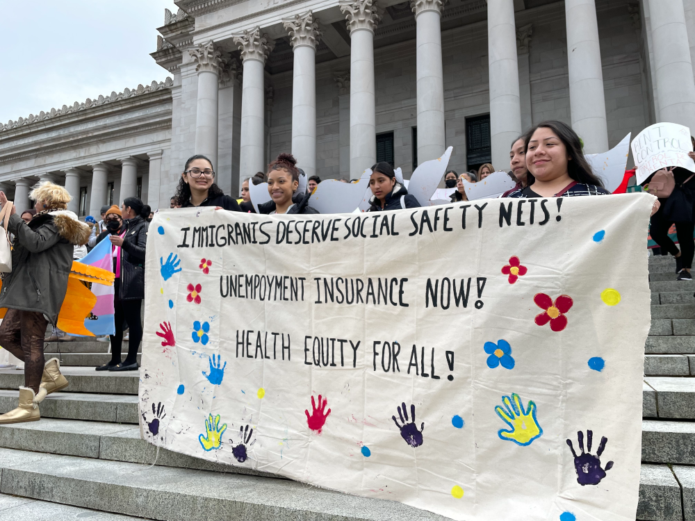 Advocates hold a banner reading "Immigrants deserve social safety nets! Unemployment insurance now! Health Equity for all!" on the steps of the State Capitol Building in Olympia